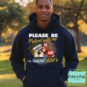 May include: Navy blue hoodie with the text "PLEASE BE Patient with me I'M FROM THE 1900's". The graphic includes a cassette tape, a retro camera, and a pink roller skate. The hoodie has a front pocket.