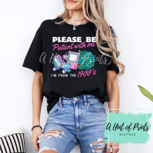May include: Black t-shirt with the text "PLEASE BE PATIENT WITH ME I'M FROM THE 1900's" in pink and white. The graphic features a roller skate, a video game console, and a disco ball.