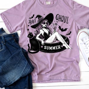 May include: A purple t-shirt with a graphic of a witch wearing a witch hat and sunglasses, floating in a pool with a black cat. The text "Hot Ghoul Summer" is printed on the shirt.