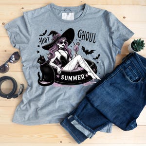 May include: Gray t-shirt with a graphic of a witch in a pool float with a black cat. The text "Hot Ghoul Summer" is printed on the shirt.