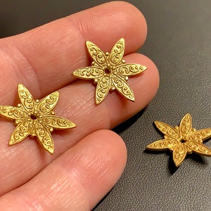 May include: Three gold-toned metal flower-shaped embellishments with intricate designs. Each flower has six points and a small hole in the center.