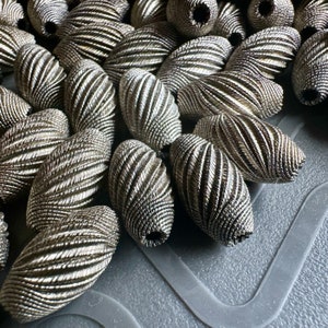 Peut inclure: Un ensemble de perles ovales torsadées de couleur argentée. Les perles ont un aspect texturé et tissé.