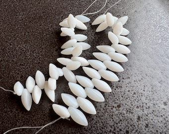 May include: White, oval-shaped beads strung on clear thread. The beads are arranged in clusters, creating a decorative pattern. The beads are smooth and have a glossy finish. The background is a dark, textured surface.