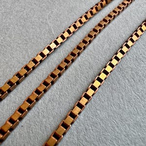 Può includere: Due collane a catena dorate con maglie quadrate. Le catene sono disposte parallelamente l'una all'altra su una superficie grigia.