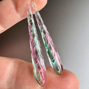 May include: A pair of long, teardrop-shaped glass earrings with a swirling pattern of pink, green, and white.