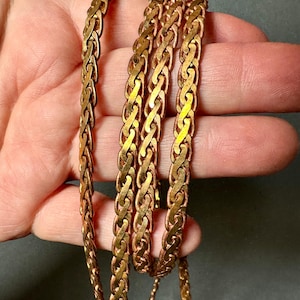 Op de afbeelding: Close-up van een koperkleurige ketting met een gevlochten ontwerp. De ketting is gemaakt van in elkaar grijpende schakels, waardoor een textuur ontstaat. De ketting heeft een warme, metallic glans en wordt gepresenteerd tegen een neutrale achtergrond.