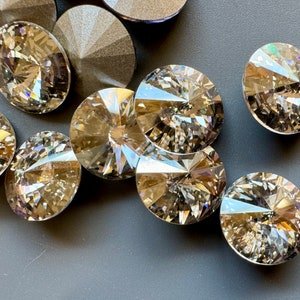May include: A group of round, clear glass rhinestones with a faceted design. The rhinestones have a light brown colour and are scattered on a grey surface.