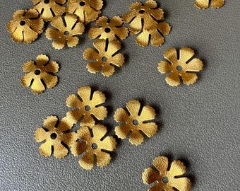 Puede incluir: Una colección de pequeños componentes metálicos dorados con forma de flor. Cada componente tiene un agujero central y cortes en forma de pétalo alrededor del borde. Los componentes están dispersos sobre una superficie gris.