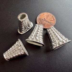 May include: Close-up of silver-colored cone-shaped beads with decorative patterns. The beads are arranged on a dark surface, next to a penny. The beads have a flared base and a detailed design of circles and lines.