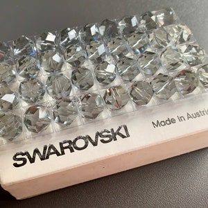 Könnte beinhalten: Eine transparente Plastikbox gefüllt mit kleinen, klaren, facettierten Glasperlen. Die Box ist mit "SWAROVSKI" und "Made in Austria" beschriftet.