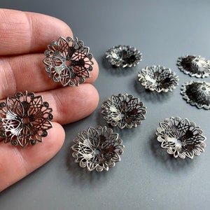 Puede incluir: Ocho adornos de metal plateado filigranado con un diseño floral. Los adornos están dispuestos sobre una superficie gris.