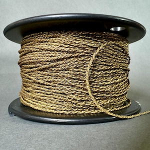 Op de afbeelding: Een zwarte houten spoel gevuld met goudkleurig koord. Het koord is strak gewikkeld en heeft een gedraaide textuur en een metallic glans. De spoel ligt op een grijze ondergrond.