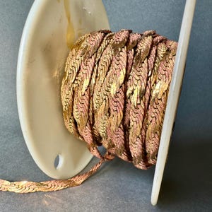 May include: A spool of gold and rose gold braided trim. The trim is a woven chain-like design.