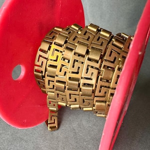 May include: A gold-colored metal chain with a repeating geometric pattern. The chain is coiled and wrapped around a red circular object.