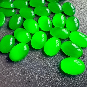May include: A collection of bright green, oval-shaped gemstones. The polished stones are scattered across a dark surface, each reflecting light. The gemstones have a smooth, glossy appearance, and vary slightly in size.