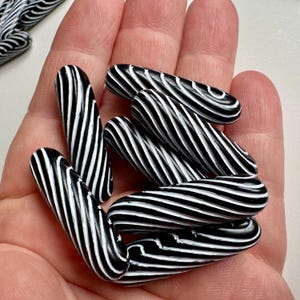 May include: A collection of black and white striped beads, held in a hand. The beads are cylindrical with a glossy finish. The stripes are evenly spaced and create a swirling pattern. The beads are a striking contrast of black and white.