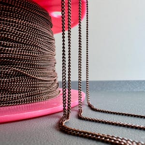 Könnte beinhalten: Eine Spule mit Kupferkette, die um eine rosa Kunststoffspule gewickelt ist. Die Kette hängt in vier Strängen herunter und zeigt die Textur und Farbe der Kette.