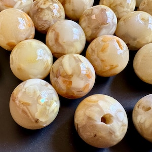 May include: A collection of round, glossy beads in shades of cream, beige, and brown. Some beads have a hole drilled through them. The beads have a marbled appearance with flecks of color, suggesting a natural material.
