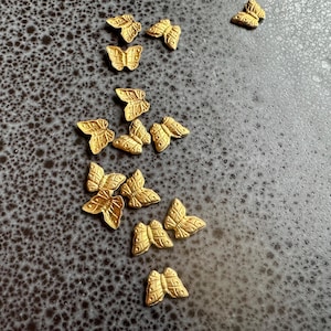 Puede incluir: Una colección de abalorios de mariposa de metal dorado. Los abalorios están esparcidos sobre una superficie gris texturizada.
