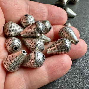 May include: A collection of silver teardrop-shaped beads with a textured, patterned design. The beads are arranged in a hand, with some beads overlapping.