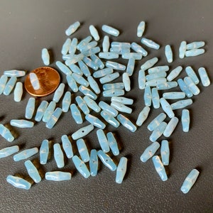 May include: A large pile of light blue glass beads, each bead is shaped like a long oval with a hole in the center. The beads are scattered on a gray surface.