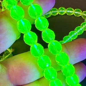 May include: A strand of faceted, translucent beads that glow with a vibrant, neon green color under ultraviolet light. The beads are spherical with multiple flat surfaces, creating a reflective, geometric appearance along the continuous string.