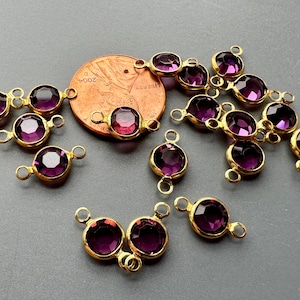 May include: A group of 15 gold-tone metal charms with purple glass stones. Each charm has two jump rings for attaching to jewelry or other crafts.