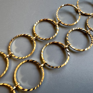 May include: Gold-toned chain necklace featuring a series of interconnected, textured oval links. The links have a diagonal striped pattern, creating a reflective surface. The necklace is displayed against a gray background.