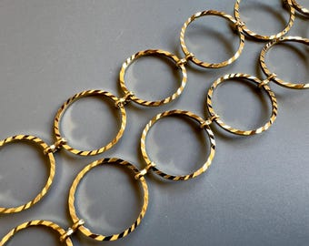 May include: Gold-toned chain necklace featuring a series of interconnected, textured oval links. The links have a diagonal striped pattern, creating a reflective surface. The necklace is displayed against a gray background.