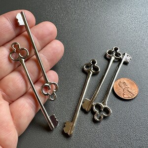 May include: Five silver-toned skeleton keys with ornate clover-shaped bowls. The keys are arranged on a hand, with one key resting on a penny.