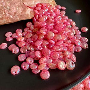 May include: A pile of iridescent pink beads spills from a brown paper bag onto a black plate. The beads are oval-shaped with a hole for stringing. The beads have a shiny, translucent appearance.