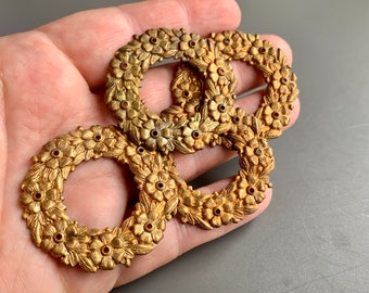 Könnte beinhalten: Vier goldfarbene Metallkränze mit floralem Design und roten Akzenten. Die Kränze liegen auf einer Hand.
