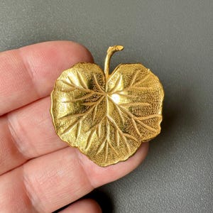 Puede incluir: Broche dorado en forma de hoja. El broche tiene vetas detalladas y una superficie texturizada. El tallo se curva ligeramente hacia arriba. El broche es probablemente de metal y es una pieza vintage o antigua.
