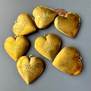 Peut inclure: Six médaillons en forme de cœur dorés avec l'inscription "I Love You" sur chacun. Les médaillons sont disposés en demi-cercle sur une surface grise.