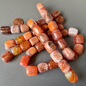 May include: A collection of orange and brown agate beads with a polished, glossy finish. The beads are rectangular with rounded edges and feature unique patterns. Some beads are strung together, while others are loose.
