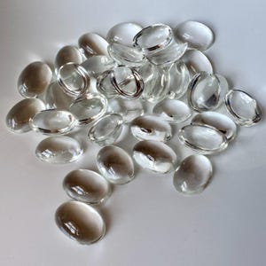 May include: A collection of clear, oval-shaped glass stones. Some stones have a silver-colored metal ring around the edge. The stones are arranged on a white surface, creating a reflective and luminous effect.