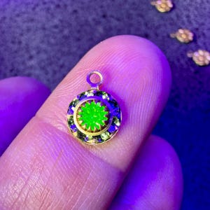 Puede incluir: Un pequeño colgante circular con una piedra central de color verde neón vibrante rodeada por un anillo de piedras facetadas de color verde más oscuro. El colgante tiene un engaste de metal dorado con una pequeña anilla circular arriba.