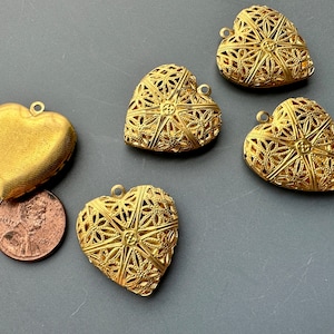 Peut inclure: Cinq pendentifs en forme de cœur de couleur or avec des motifs de filigrane complexes. Quatre des pendentifs ont des motifs ajourés, tandis qu'un est plein. Les pendentifs sont disposés sur une surface grise.