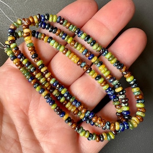 Puede incluir: Hebras de pequeñas cuentas de colores en tonos verdes, marrones, amarillos y azules. Las cuentas están ensartadas, creando un collar o pulsera. Las cuentas se sostienen en una mano.