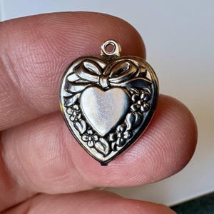 May include: Silver heart-shaped pendant with a bow and floral design. The pendant features a central heart shape and is surrounded by delicate flowers. The pendant has a small loop at the top for attaching to a chain or bracelet.