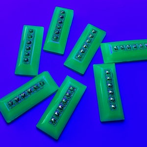 Può includere: Sette bottoni rettangolari in vetro verde con dettagli argentati e strass. I bottoni sono su uno sfondo blu.