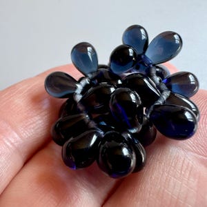 May include: A cluster of dark blue glass beads in the shape of teardrops, arranged in a circular pattern. The beads are strung together with a thin, clear thread.