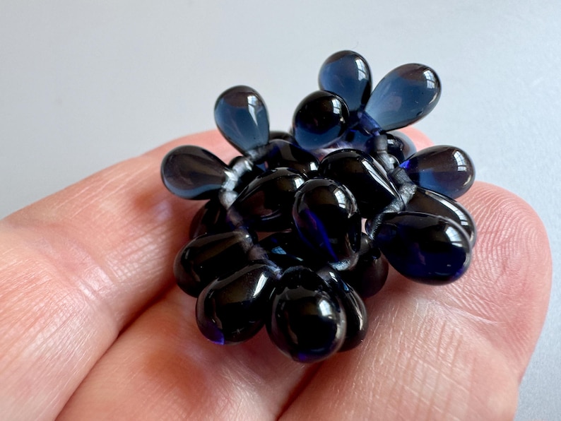 May include: A cluster of dark blue glass beads in the shape of teardrops, arranged in a circular pattern. The beads are strung together with a thin, clear thread.