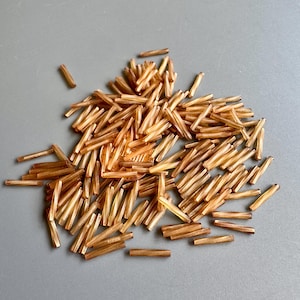Könnte beinhalten: Ein Haufen kleiner, goldbrauner Glasperlen, jede zylindrisch mit facettierten Kanten. Die Perlen sind auf einer grauen Oberfläche verstreut, wodurch ein strukturiertes Aussehen entsteht. Ideal für die Schmuckherstellung und das Basteln.