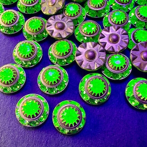 Puede incluir: Una colección de botones circulares con una piedra central facetada de color verde neón brillante rodeada por un engaste metálico tipo engranaje. Algunos botones muestran una parte superior metálica en forma de estrella, otros la piedra.