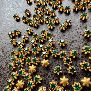 May include: A collection of small, gold-colored metal embellishments with emerald green gemstones. The embellishments are scattered across a dark surface, with some in a star shape. These decorative elements are ideal for jewelry making or craft projects.