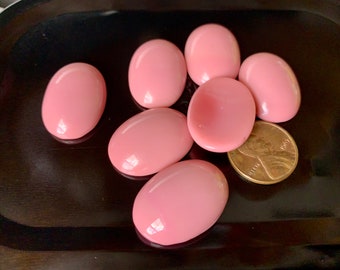 Könnte beinhalten: Sieben ovale, rosafarbene Cabochons, einige mit einer leichten Kuppelform, liegen auf einer schwarzen Oberfläche.