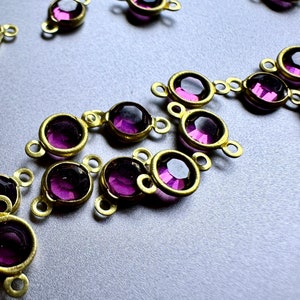 May include: A group of purple glass beads with gold metal settings. The beads are arranged in a loose pattern on a silver surface.