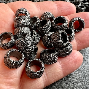 Puede incluir: Un montón de pequeños anillos de metal negro con una superficie texturizada. Los anillos tienen un agujero en el centro y miden aproximadamente 2,5 cm de diámetro.