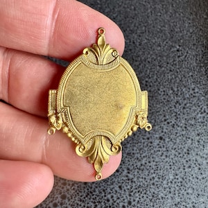 May include: Ornate gold-colored metal pendant with an oval center and decorative filigree edges. The pendant has a small loop at the top for hanging and is likely vintage or antique. The design includes floral and scroll elements.
